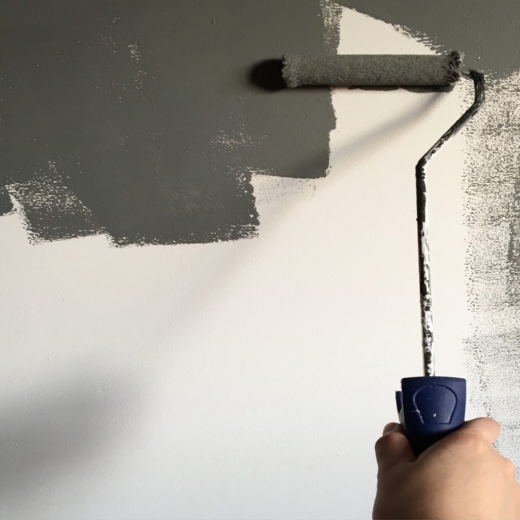 A light-skinned hand holds up a paint roller in front of a wall that is half grey, half white.