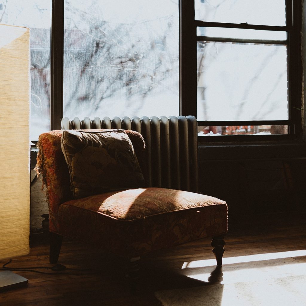 A chair and a floor lamp sitting beside a window are illuminated by the sun.