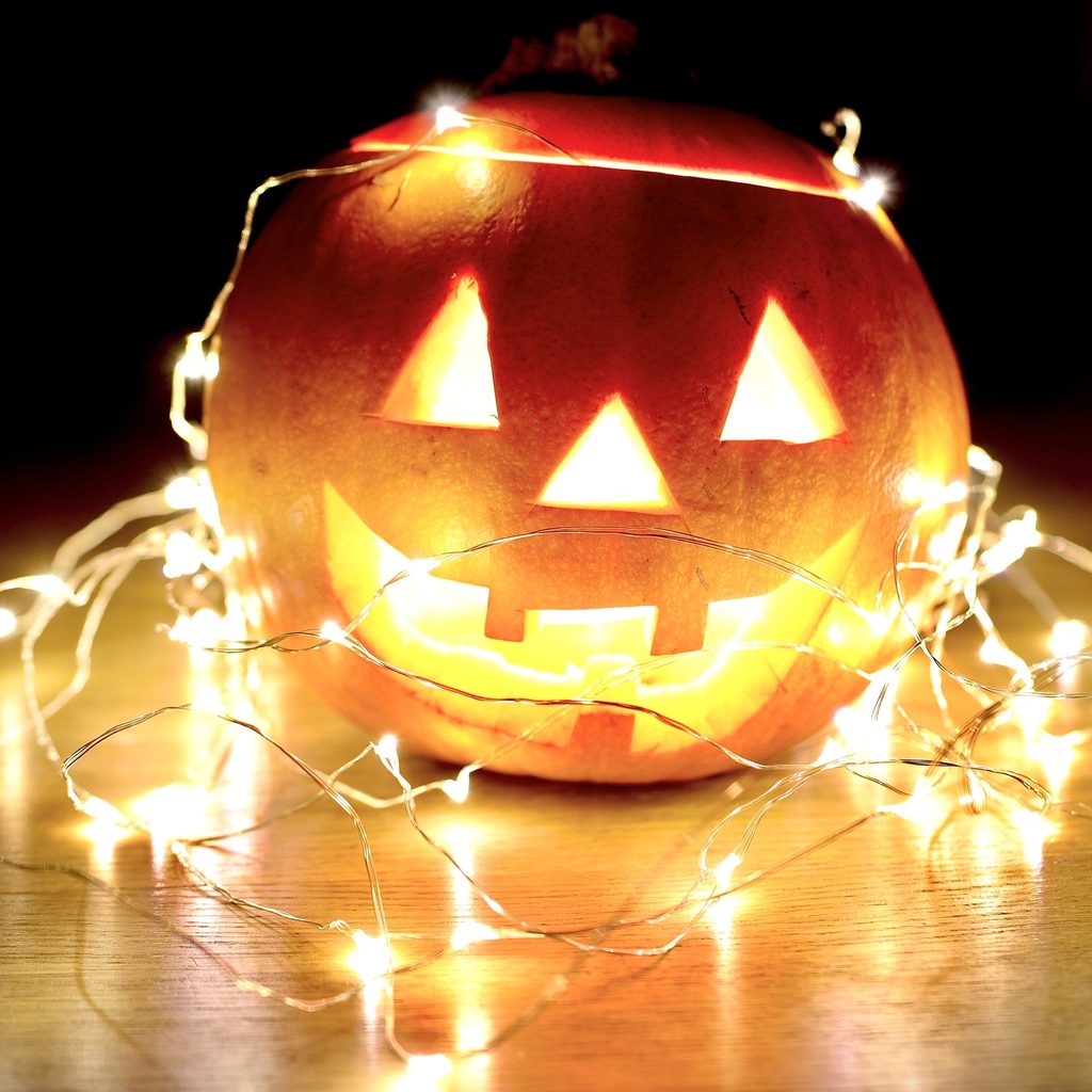 A orange pumpkin sits atop a light wooden surface covered by fairy lights and lit from within
