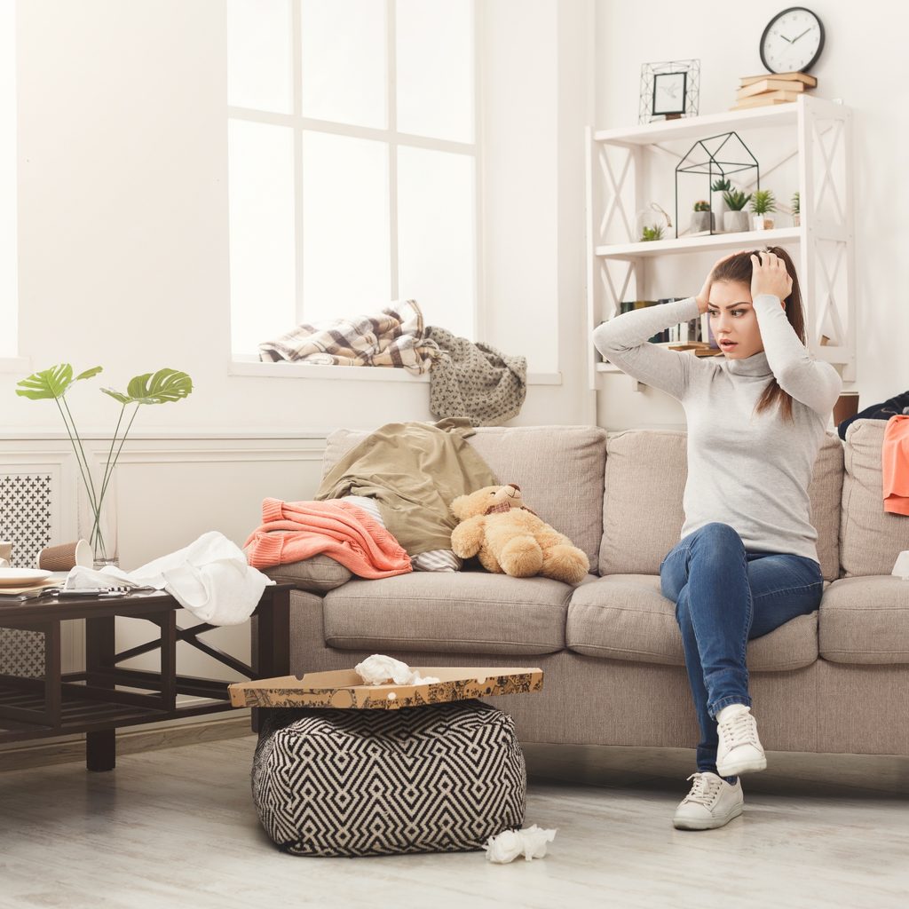 Distressed girl sitting on couch surrounded by clutter.