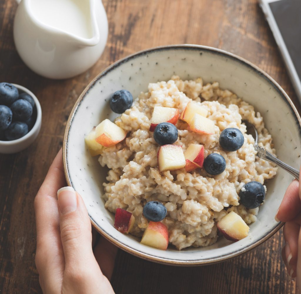 Person with bowl of oatmeal with blue berries and apples in it.
