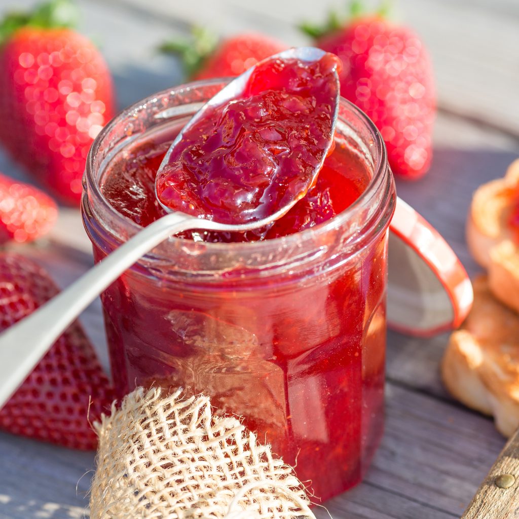 Spoon scooping strawberry jam out of a jar with toast and fresh strawberries on the table around it