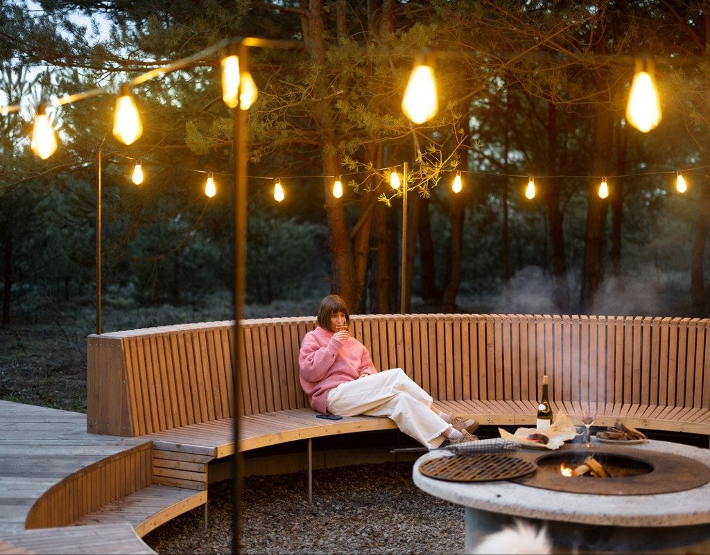 Young woman sits on round bench while resting at beautiful bbq area illuminated with garlands in forest at dusk