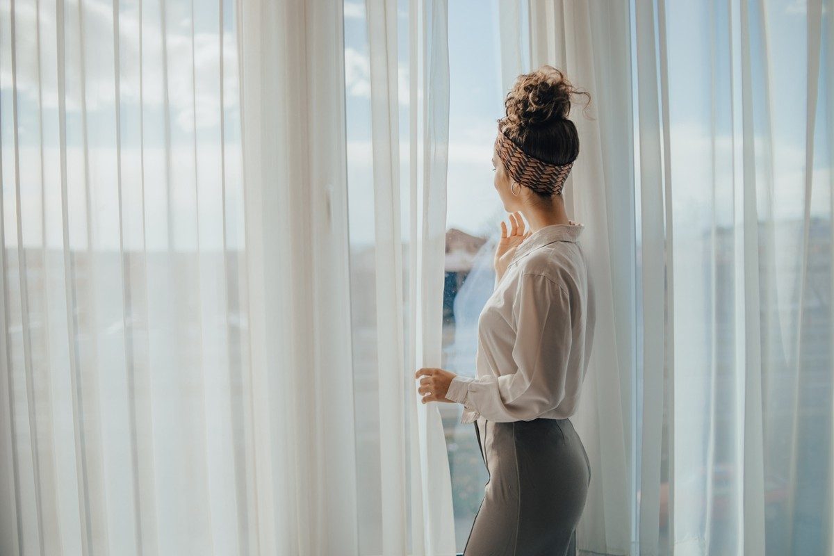 One young woman opens the curtains in the room in the morning at sunrise, she is dressed in a white shirt, ready for work and daily duties, morning routine