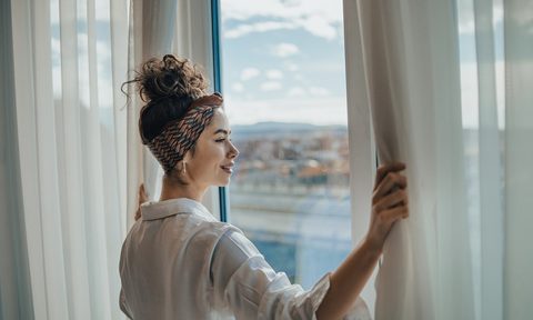 One young woman opens the curtains in the room in the morning at sunrise, she is dressed in a white shirt, ready for work and daily duties, morning routine