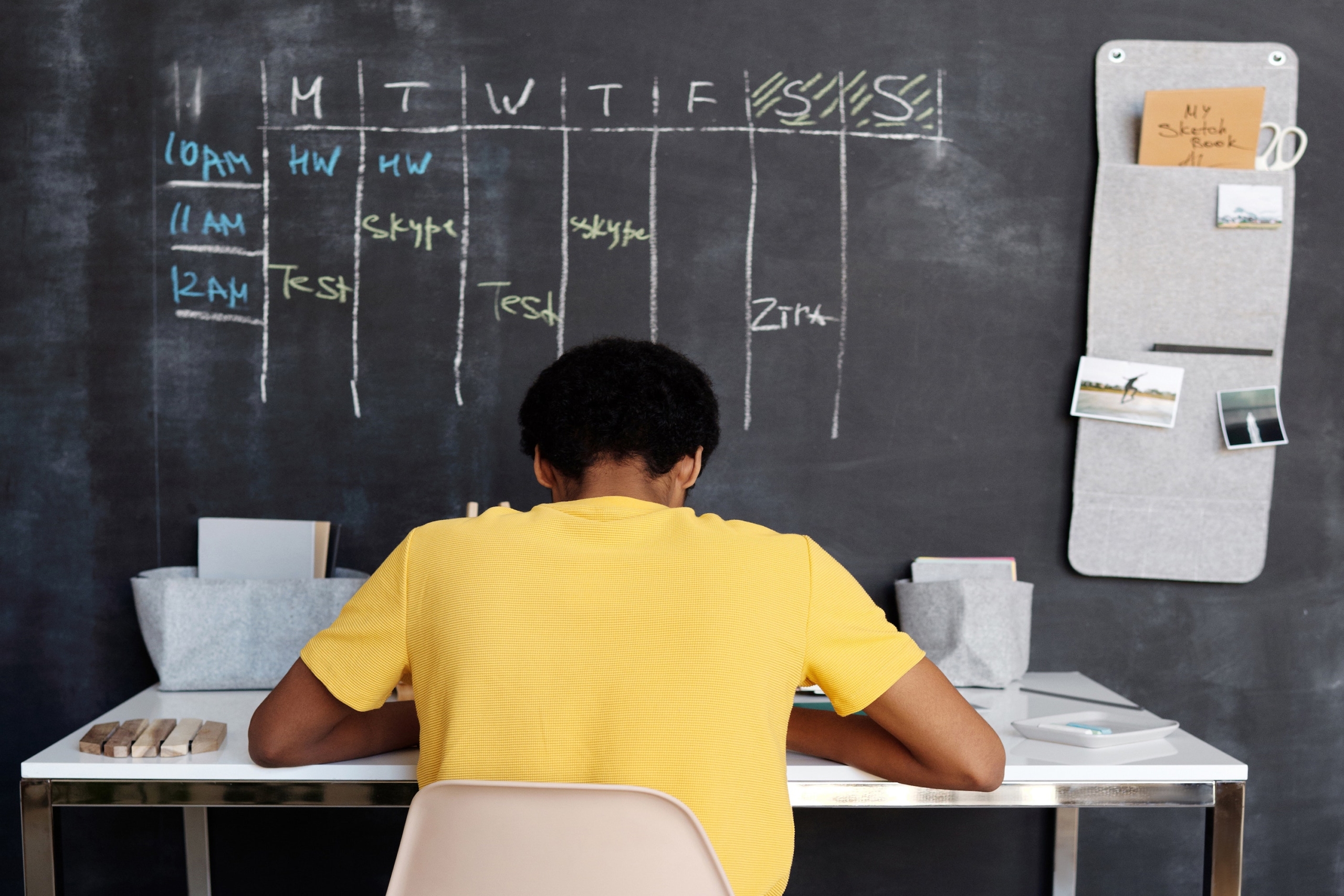A young boy in a yellow shirt studies at a desk placed in front of a black chalkboard wall
