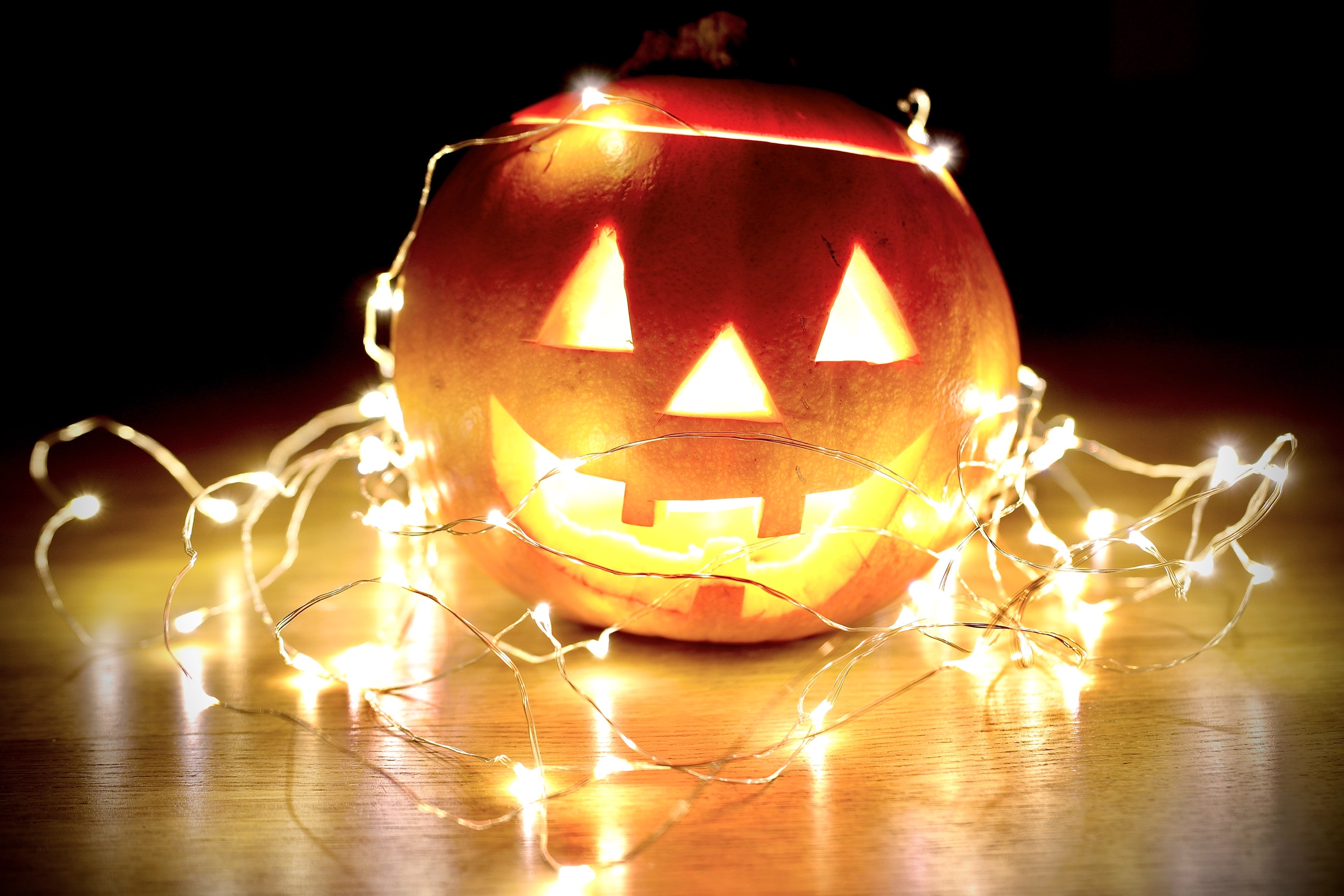 A orange pumpkin sits atop a light wooden surface covered by fairy lights and lit from within
