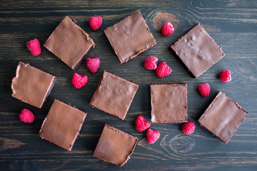Individual brownie squares on a wood surface surrounded by raspberries