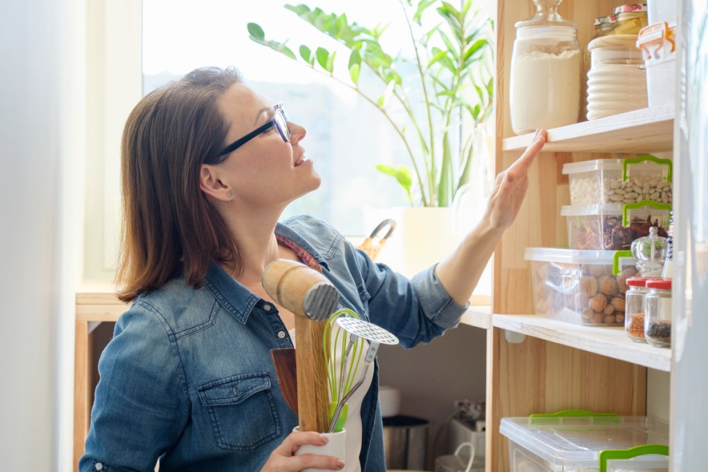 Woman with cooking accessories in kitchen interior, background wooden shelf in pantry with utensils, food cans