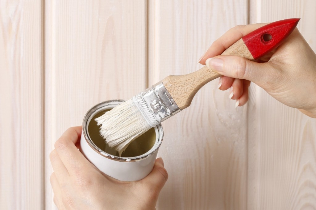 Person holding small paint can and brush in front of wood paneling