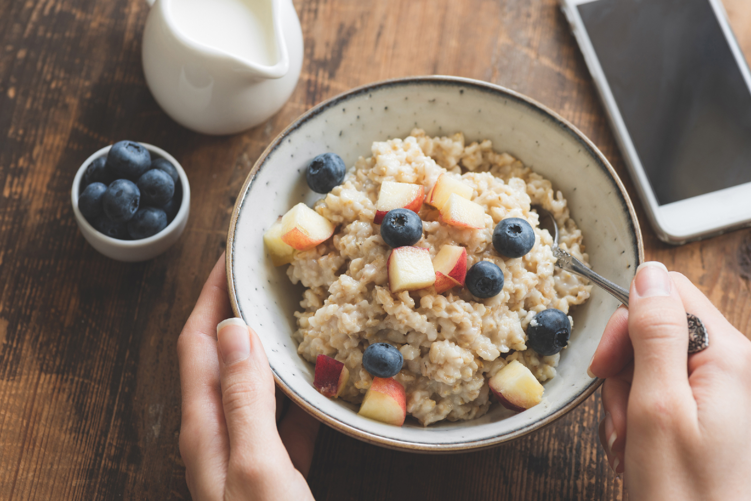 Person with bowl of oatmeal with blue berries and apples in it.