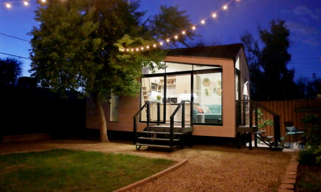 outdoor light timer benefits a tiny house with large glass windows  sits in the backyard at night surrounded by trees and par