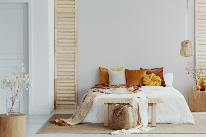 Brown and orange pillows on a white bed in natural bedroom interior with wicker lamp and wooden bedside table with vase