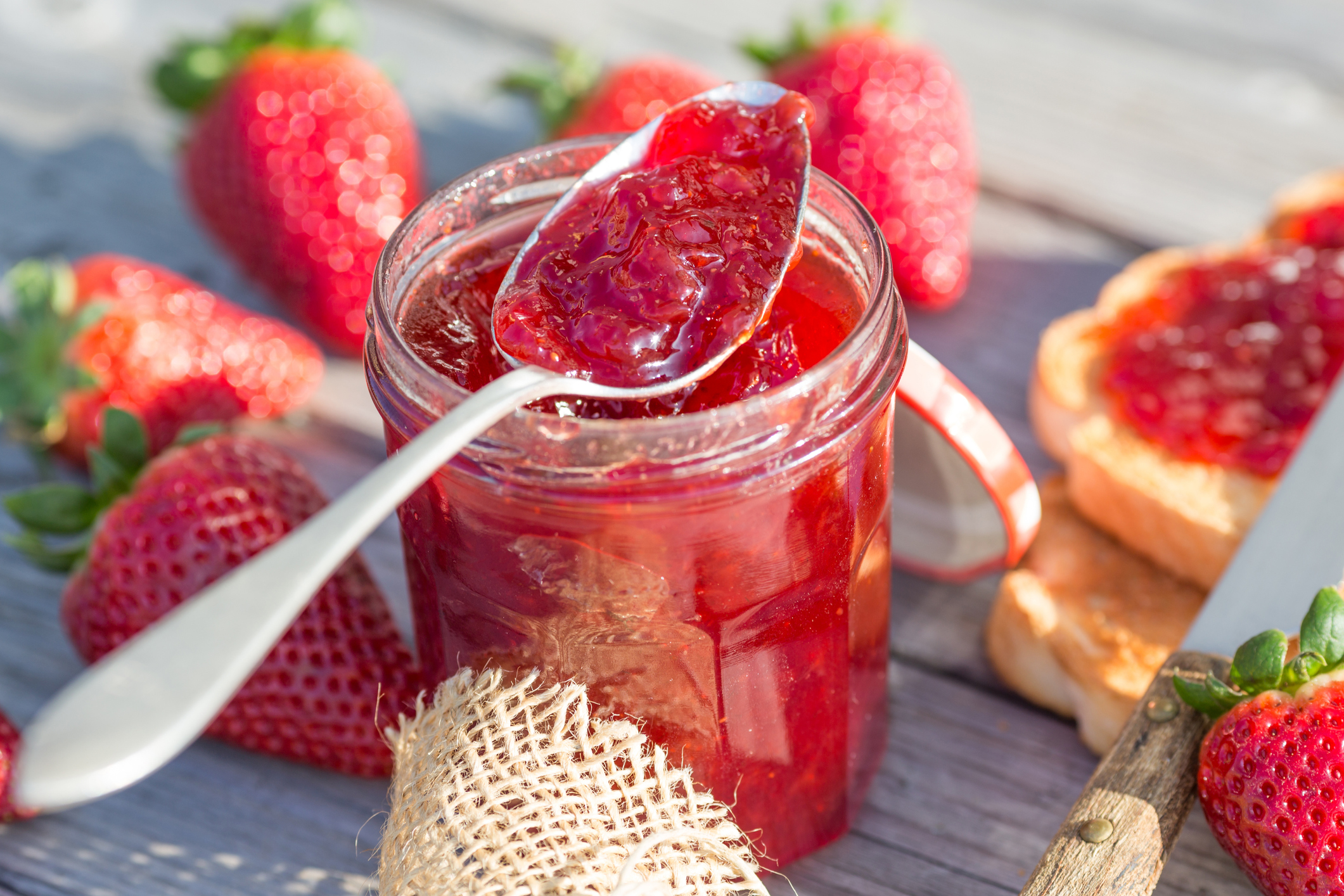 Spoon scooping strawberry jam out of a jar with toast and fresh strawberries on the table around it