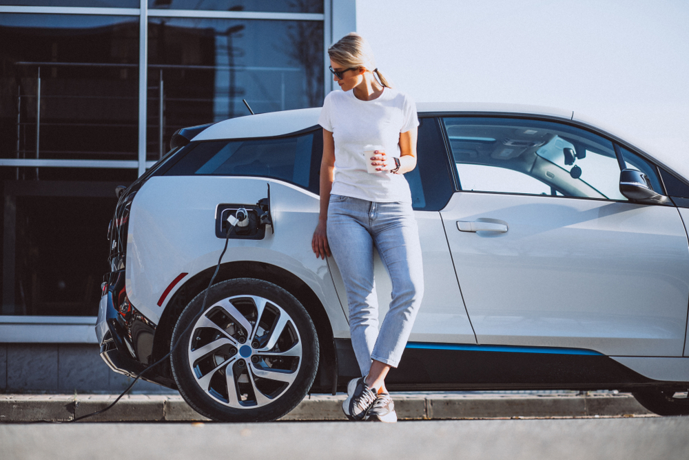 Woman holding coffee next to charging electric vehicle