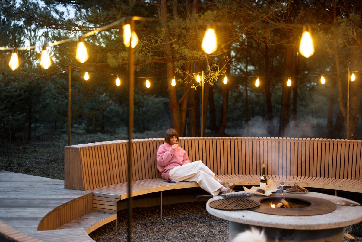 Young woman sits on round bench while resting at beautiful bbq area illuminated with garlands in forest at dusk