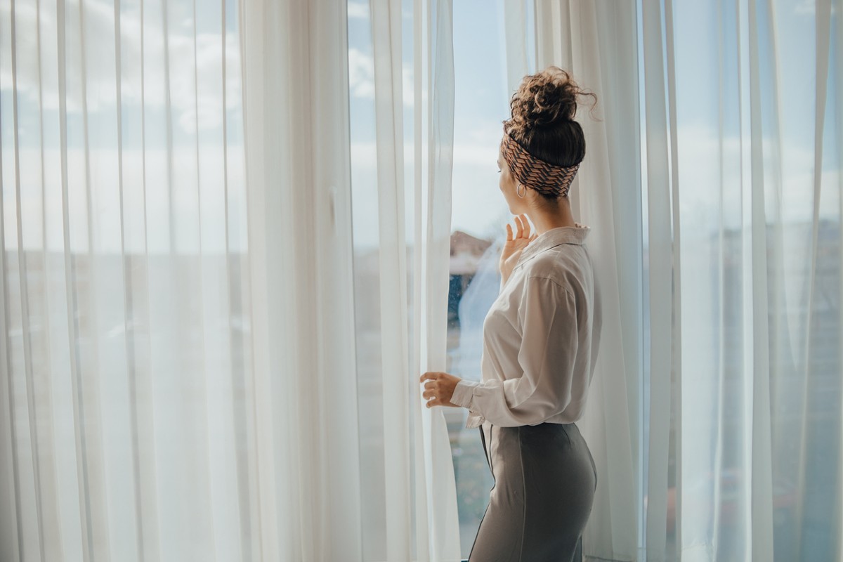 One young woman opens the curtains in the room in the morning at sunrise, she is dressed in a white shirt, ready for work and daily duties, morning routine